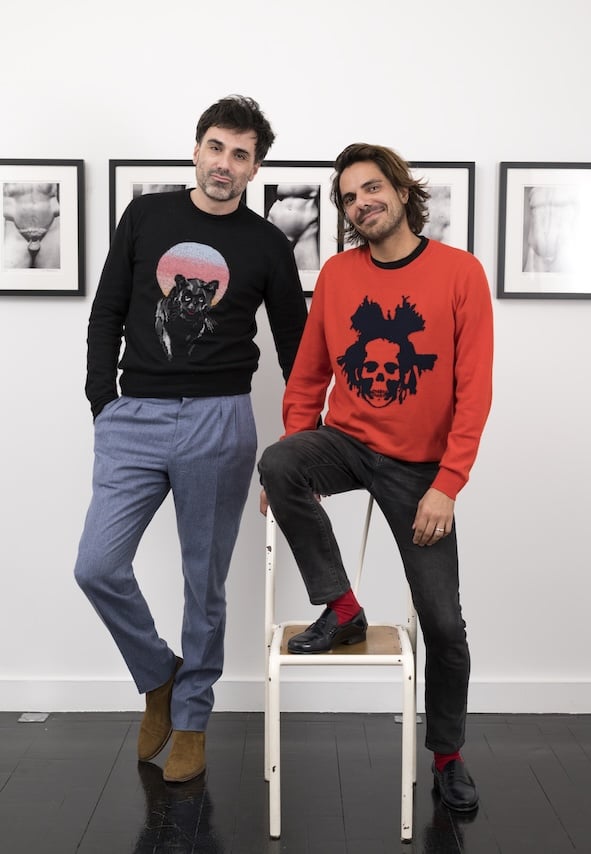 This image shows two men posing in front of a wall adorned with black-and-white photographs. The man on the left is wearing a black sweater with a graphic of a panther and light blue trousers, standing casually with his hands in his pockets. The man on the right, sitting on a chair, is wearing a red sweater with a black skull design and dark jeans. Both are dressed stylishly, with the man on the right adding a pop of color with red socks and black shoes. The mood is relaxed and artistic, with both men appearing comfortable in the gallery-like setting