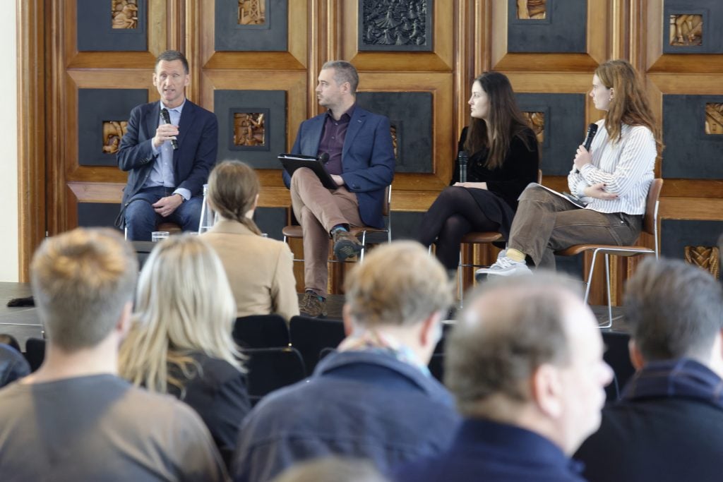 Four panelists on stage speaking at a conference with wooden decorative panels behind them.