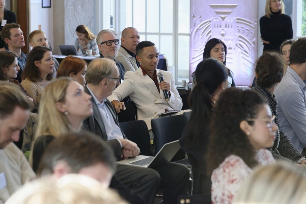 Man in white suit asking a question at a conference panel discussion.