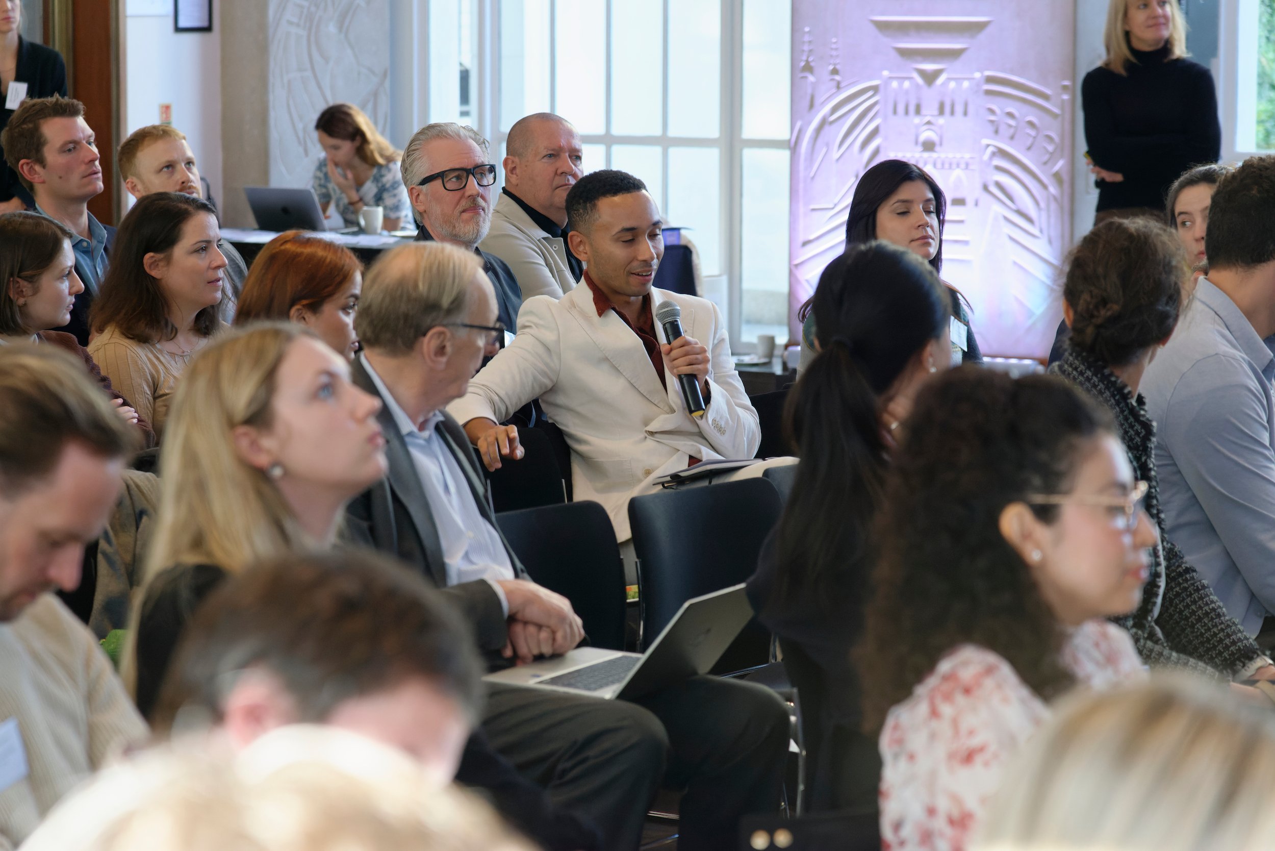 Man in white suit asking a question at a conference panel discussion.