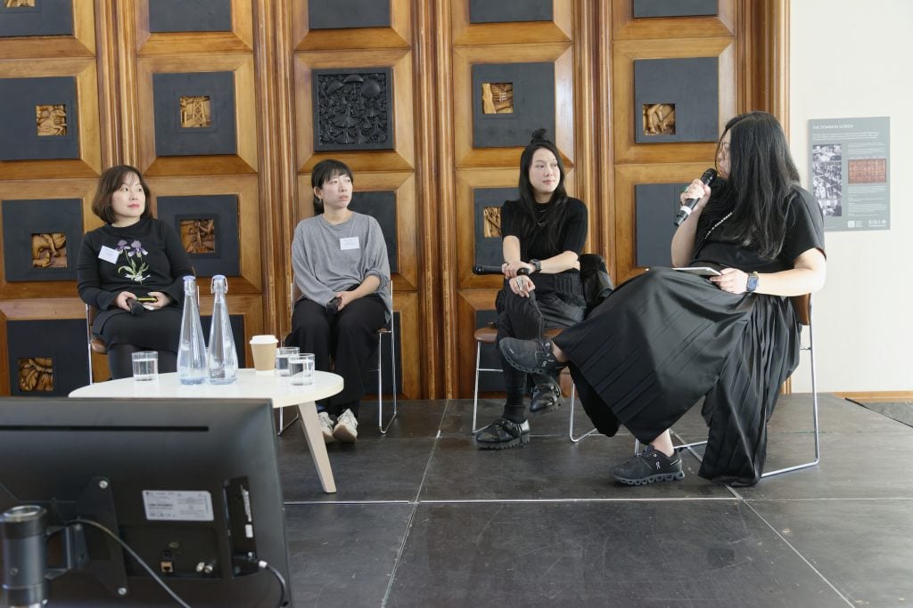 Four panelists on stage speaking at a conference with wooden decorative panels behind them.