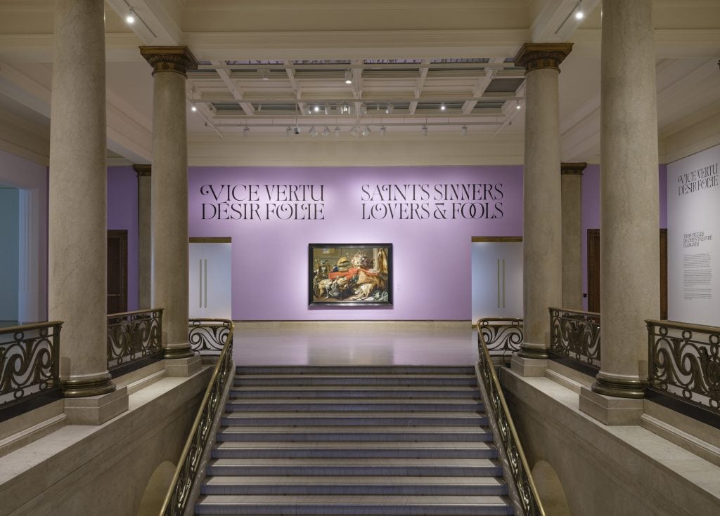 a grand staircase with four big columns and a purple wall with a big painting on it