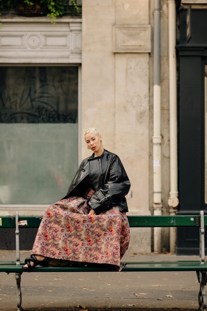 a woman in a leather jacket and a tapestry-like skirt sits a top the upper part of a public bench in paris