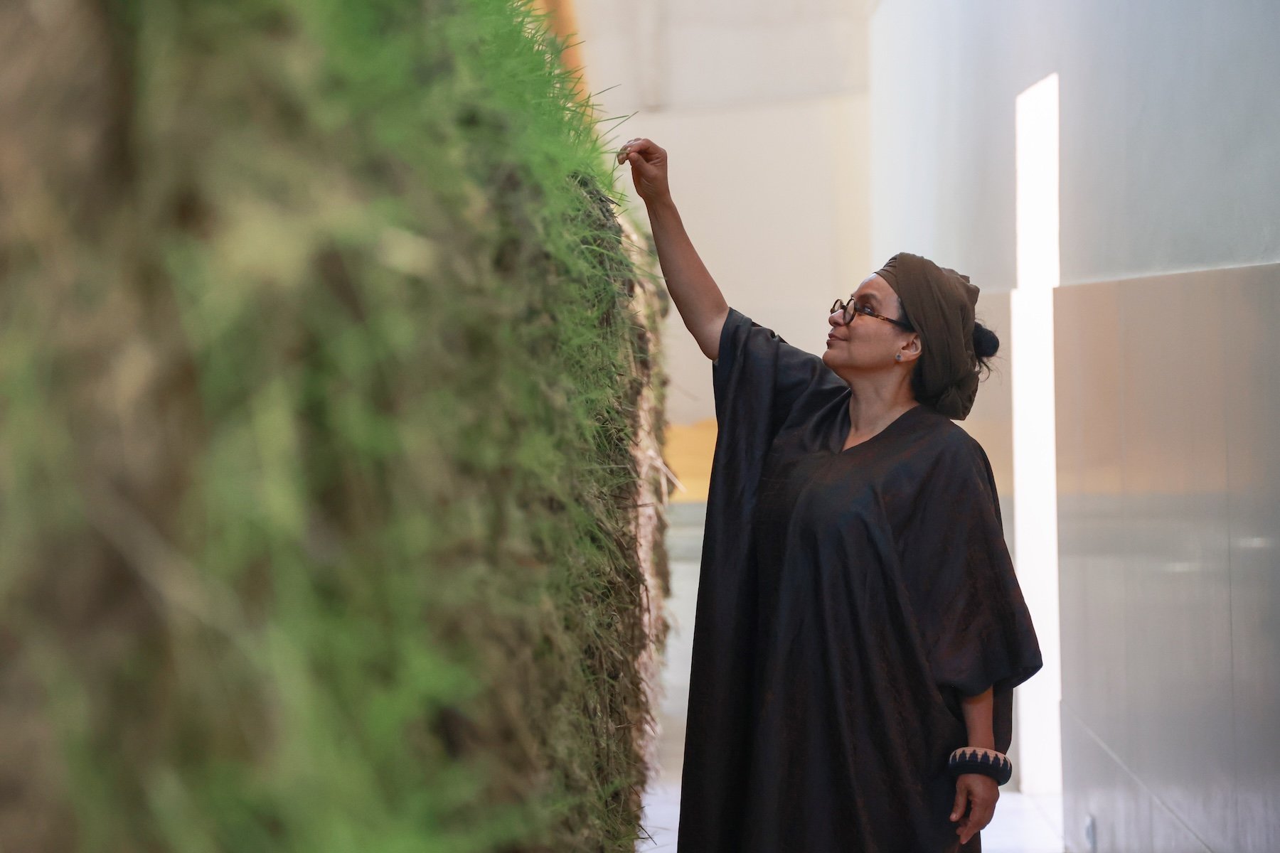 A woman in a dark robe touches a tall wall of grass or natural material, inspecting it carefully in a brightly lit, modern space