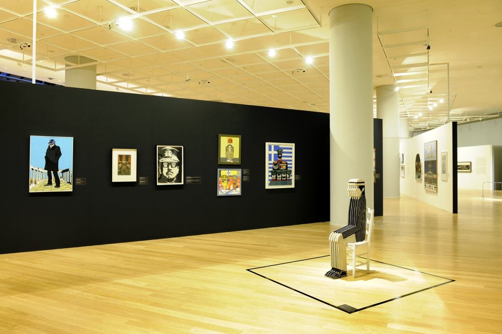 An installation view at the National Gallery - Alexandros Soutsos Museum featuring a series of framed artworks on a black wall. The displayed artworks include portraits and colorful compositions, likely reflecting social or political themes. In the foreground, there is a striped sculpture of a seated figure on a white chair, enclosed within a marked square on the wooden floor. The space has bright lighting and a clean, modern gallery layout.