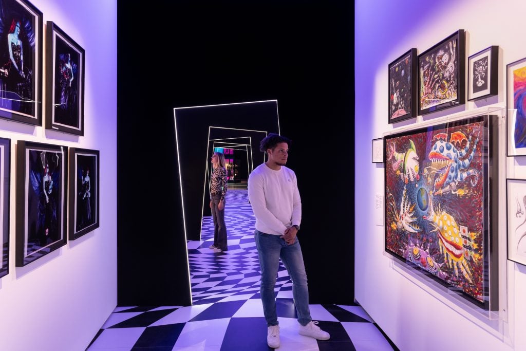 A man in white top and jeans looks at colourful paintings of monsters mounted on a white call on the left of the image, and on the right, there are colourful portraits of women. The room has black and white floor tiles and tilted door frames.