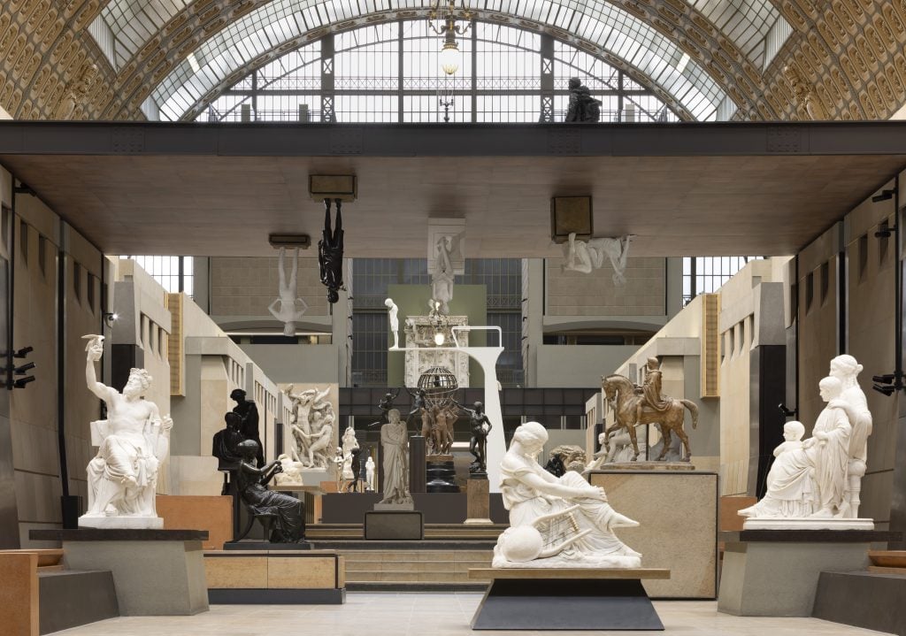 A contemporary white statue of a boy looking at his smartphone stands in the Musée d’Orsay’s grand sculpture hall. Surrounding the statue are classical bronze and marble sculptures, creating a contrast between old and new interpretations of the human form.