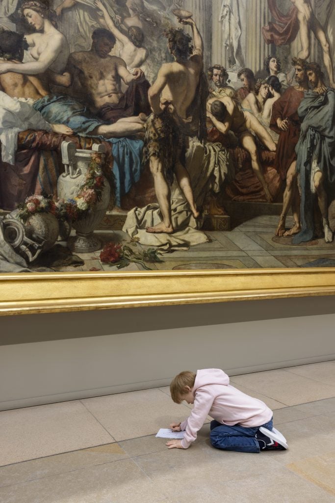 A young child sits on the floor, drawing with a pencil in front of a large neoclassical painting of a bacchanalian Roman scene in the Musée d’Orsay. The child is part of an art installation that contrasts the grand historical painting with the simple act of drawing.