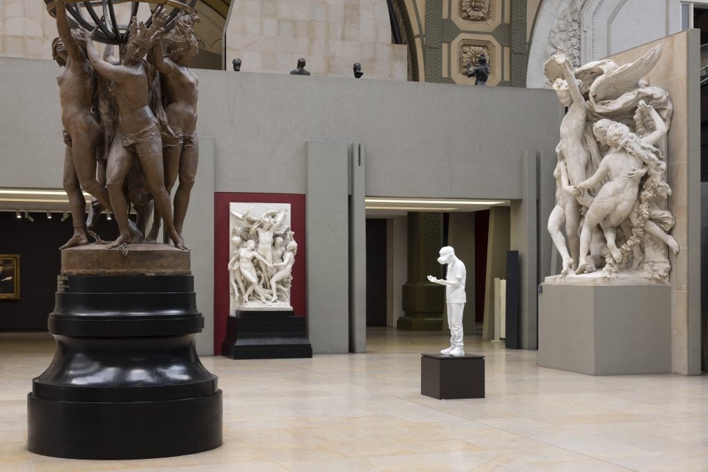 A white statue of a boy stands at the edge of a tall diving board, part of an artistic installation in the Musée d’Orsay. The space is filled with classical sculptures, while a large neoclassical painting adorns the nearby wall, creating a juxtaposition of contemporary and traditional art.