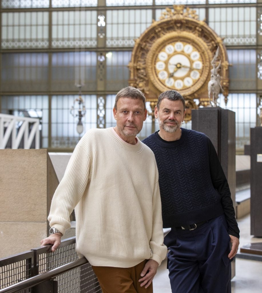 This image shows two men standing in an interior space with an elegant, large clock in the background, likely inside a museum or historical building. The man on the left wears a cream-colored ribbed sweater and brown pants, while the man on the right is dressed in a black textured sweater and navy blue trousers. They are casually posed, with the man on the left leaning slightly against a railing. The large, ornate clock behind them adds a sense of grandeur to the setting, and the overall atmosphere suggests a blend of modern style with a classical environment.