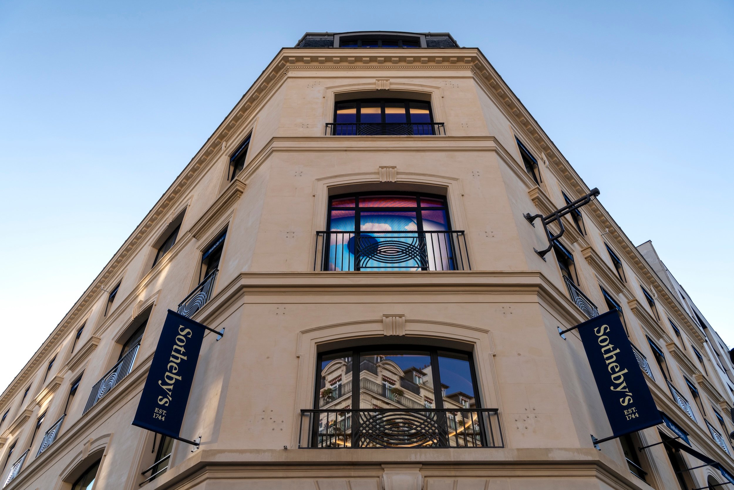 A modern photograph showing the exterior of a corner building where Sotheby's Paris is, with beige stone walls, identified by two hanging banners as Sotheby’s, established in 1744. The building has large arched windows with reflective glass, showcasing the reflection of a nearby structure. The angle of the shot looks upward, emphasizing the height and sharp angles of the building. The bright sky above creates a clean backdrop, while artistic window details and the prominent blue Sotheby’s banners add sophistication and elegance to the overall design