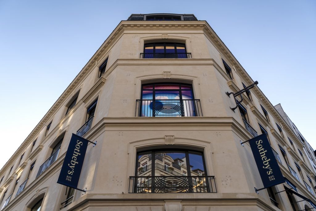 A modern photograph showing the exterior of a corner building where Sotheby's Paris is, with beige stone walls, identified by two hanging banners as Sotheby’s, established in 1744. The building has large arched windows with reflective glass, showcasing the reflection of a nearby structure. The angle of the shot looks upward, emphasizing the height and sharp angles of the building. The bright sky above creates a clean backdrop, while artistic window details and the prominent blue Sotheby’s banners add sophistication and elegance to the overall design