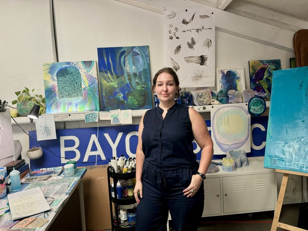 Falon Mihalic, a Houston artist who created an installation titled Abandoned Garden for Meow Wolf Radio Tave in her studio space. She is standing in front of some blue tinted paintings.