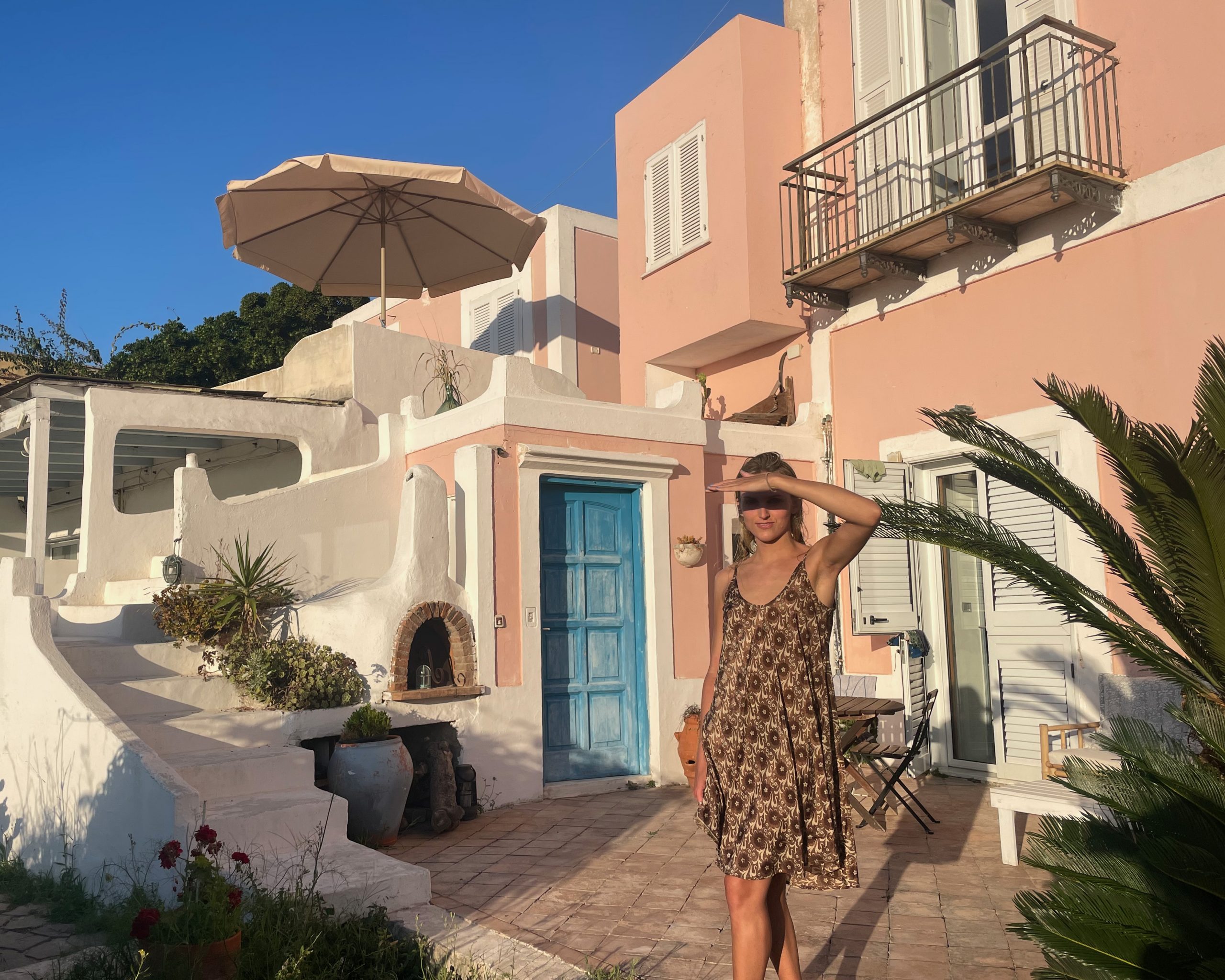 a woman in a sun dress shields her eyes from the sun in front of a mediterranean house