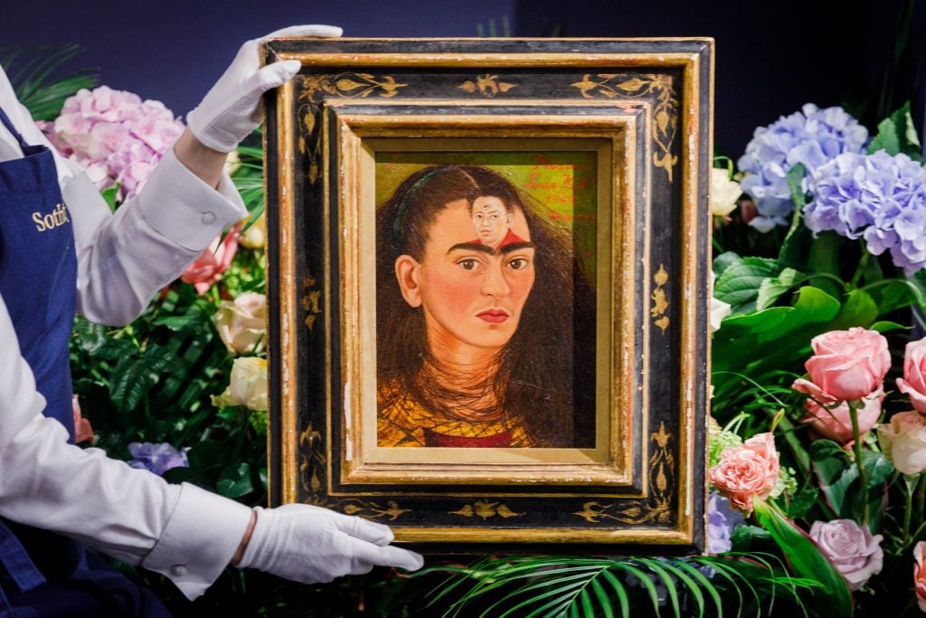 a white-gloved sotheby's employee hold a painting with a depiction of the artist Frida Kahlo. The painting of the artist show her husband, Diego Rivera, on her forehead and he has a third eye.