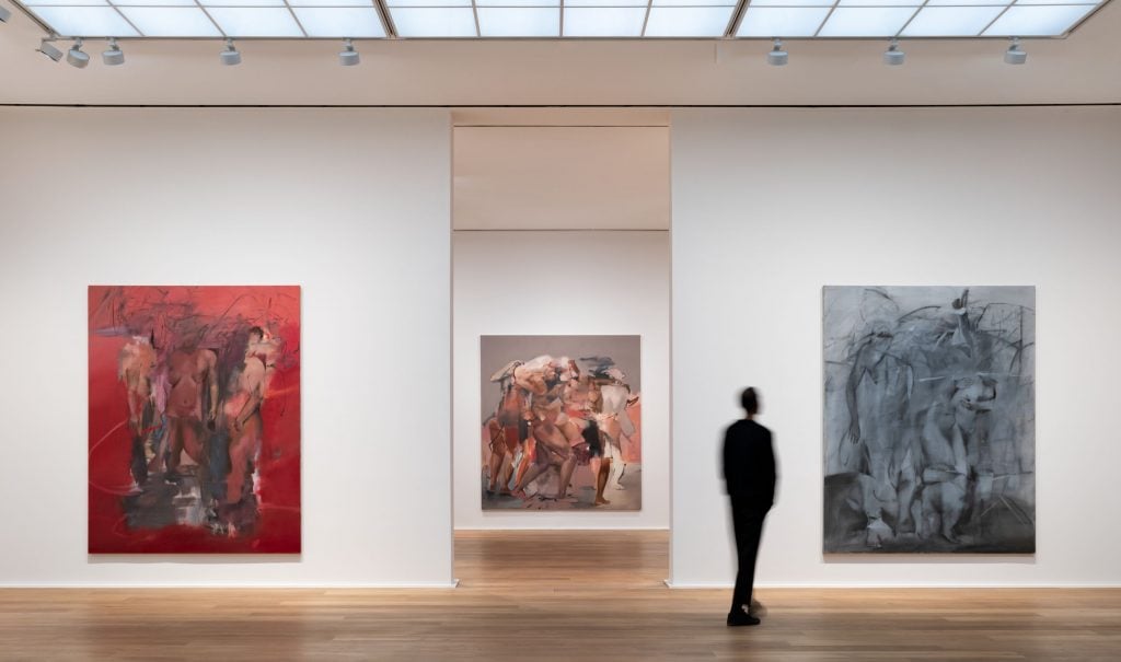 a photograph of a large, light, white-walled gallery space installed with large semi-figurative paintings. There are two on either side of open passage into a subsequent room and through the line of sight you can see a third painting in the background. The foreground two paintings are red with flesh tones on the left and silvery with monochromatic details on the right.