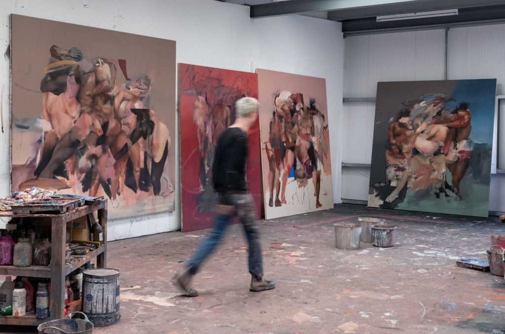 a studio space with large semi-figurative paintings hanging or leant against the walls, a slim male figure can be seen in motion walking through the space with purpose, his back turned to us