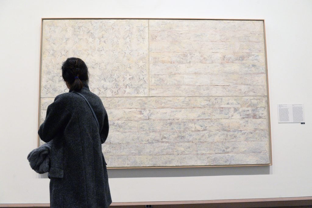 a visitor wearing all black stands in front of a painting of an american flag entirely done in shades of white