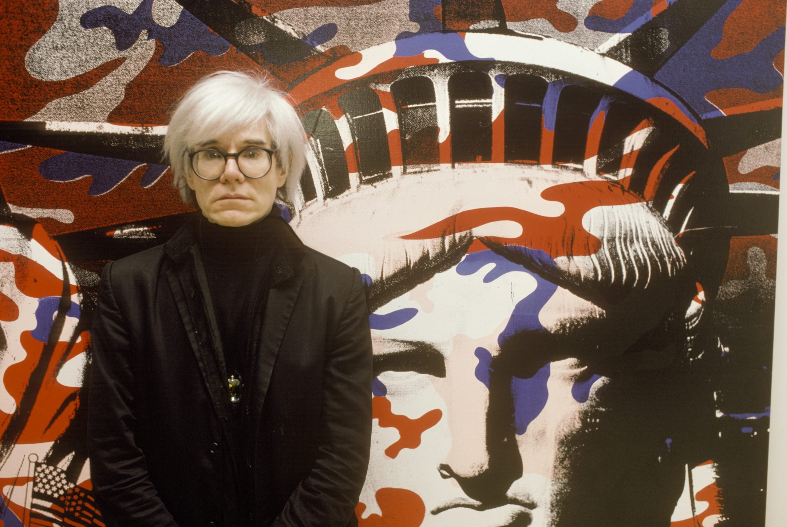 the artist andy warhol in a black jumper, glasses and white wig stands in front of a painting of the statue of liberty