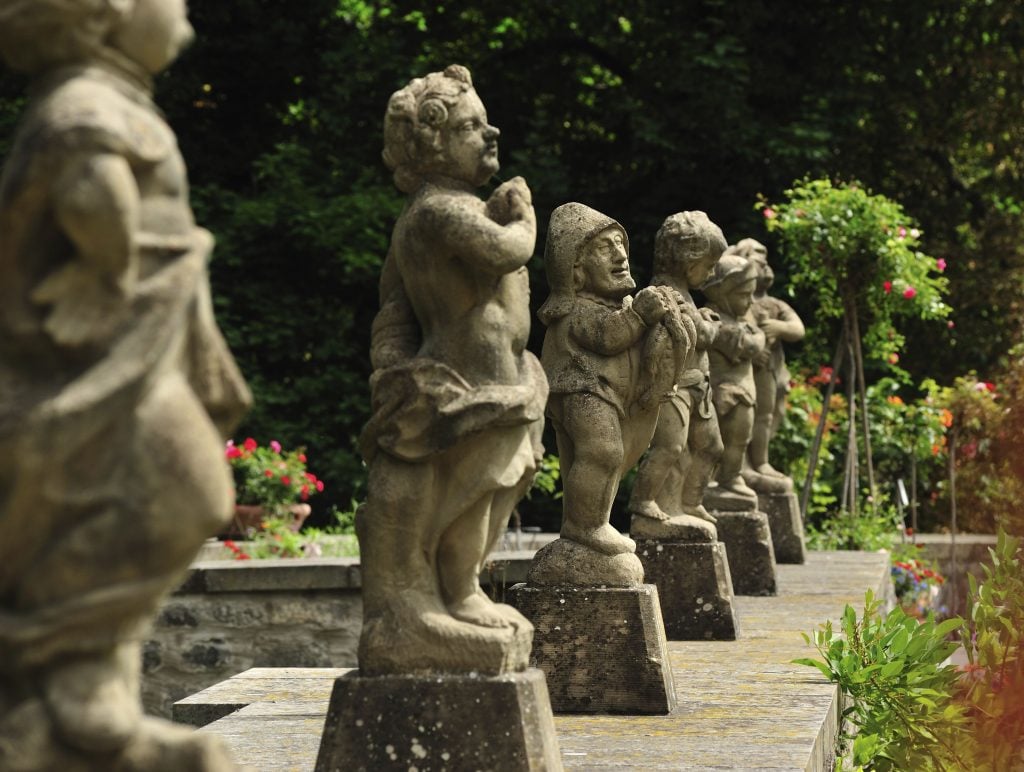 A row of stone statues depicting dwarves or small figures in various poses stands on a garden pathway, surrounded by lush greenery and vibrant flowers.