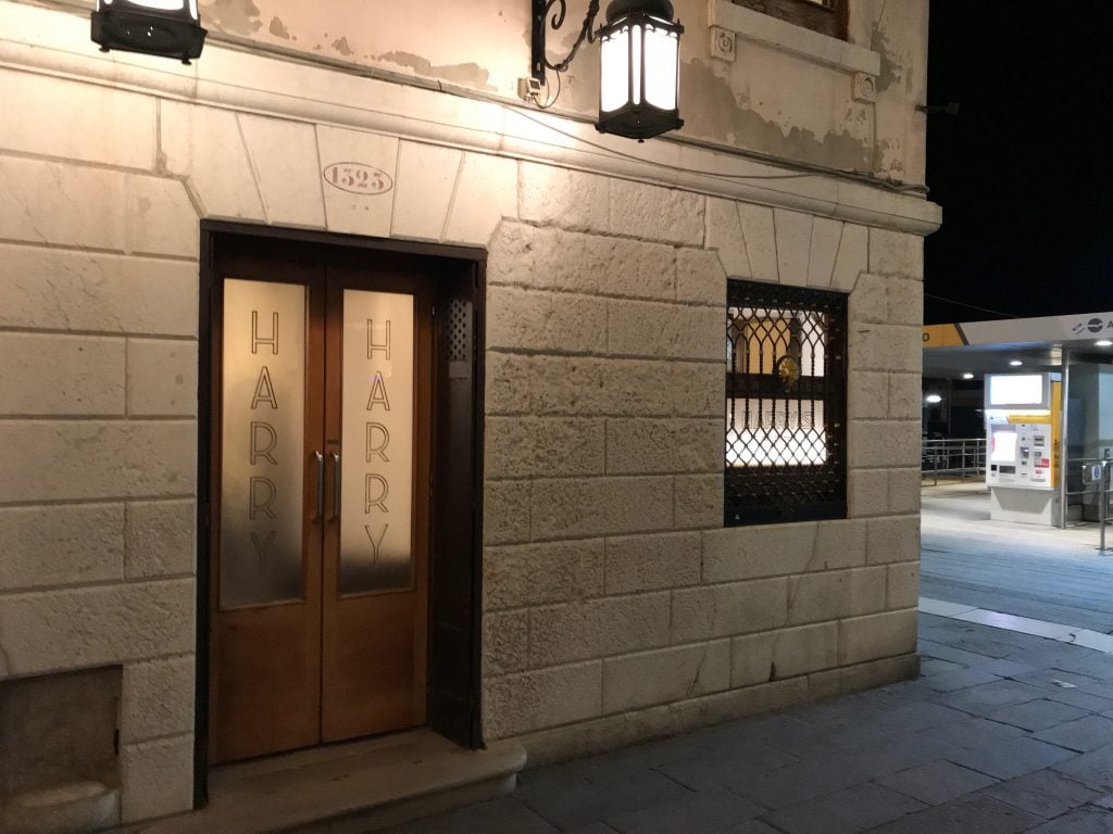 Harry's Bar in Venice, Italy, in 1998. The exterior of the bar, in an old stone building with frosted glass panels reading "Harry" in the wooden doors, is seen at night.