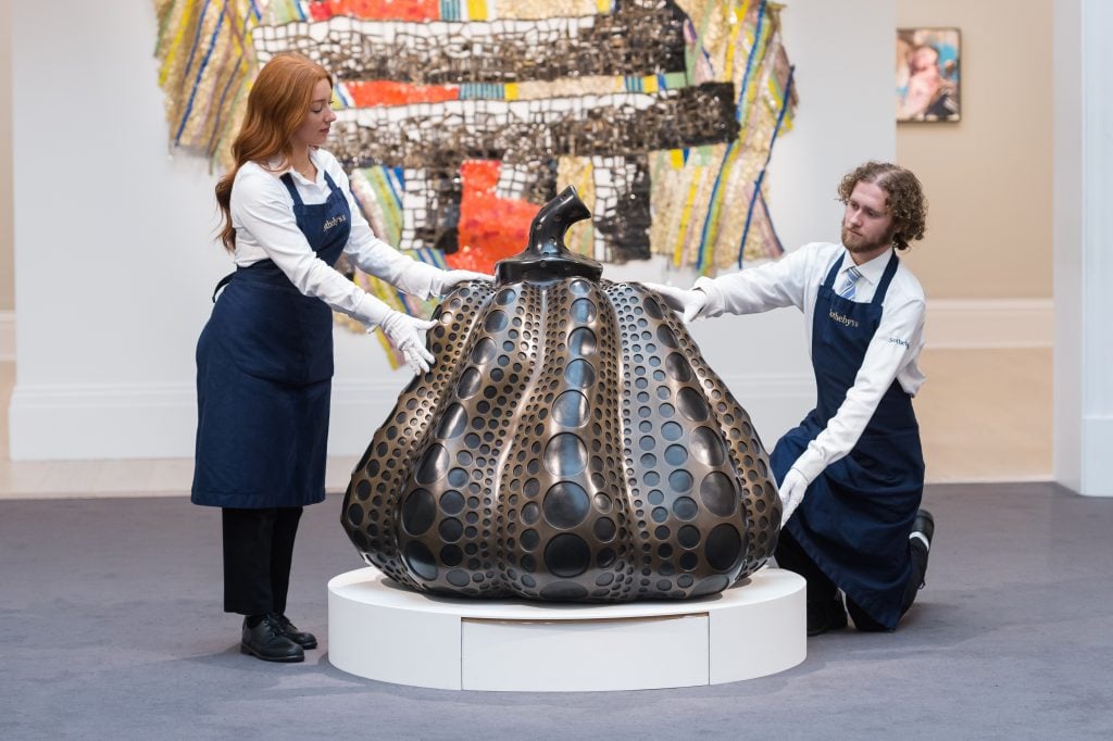 two attendants wearing blue aprons hold a bronze sculpture of a pumpkin