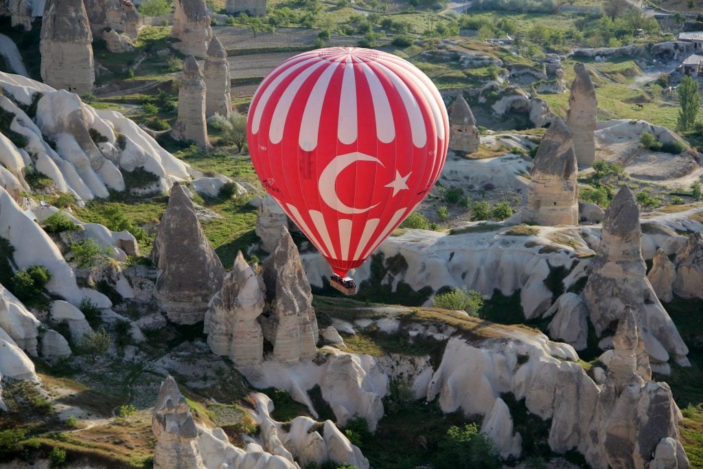 A hot air ballon in the colors and design of the Turkish flag flies over a natural landscape of earthern pillars, smooth crags, and grassy boulders.