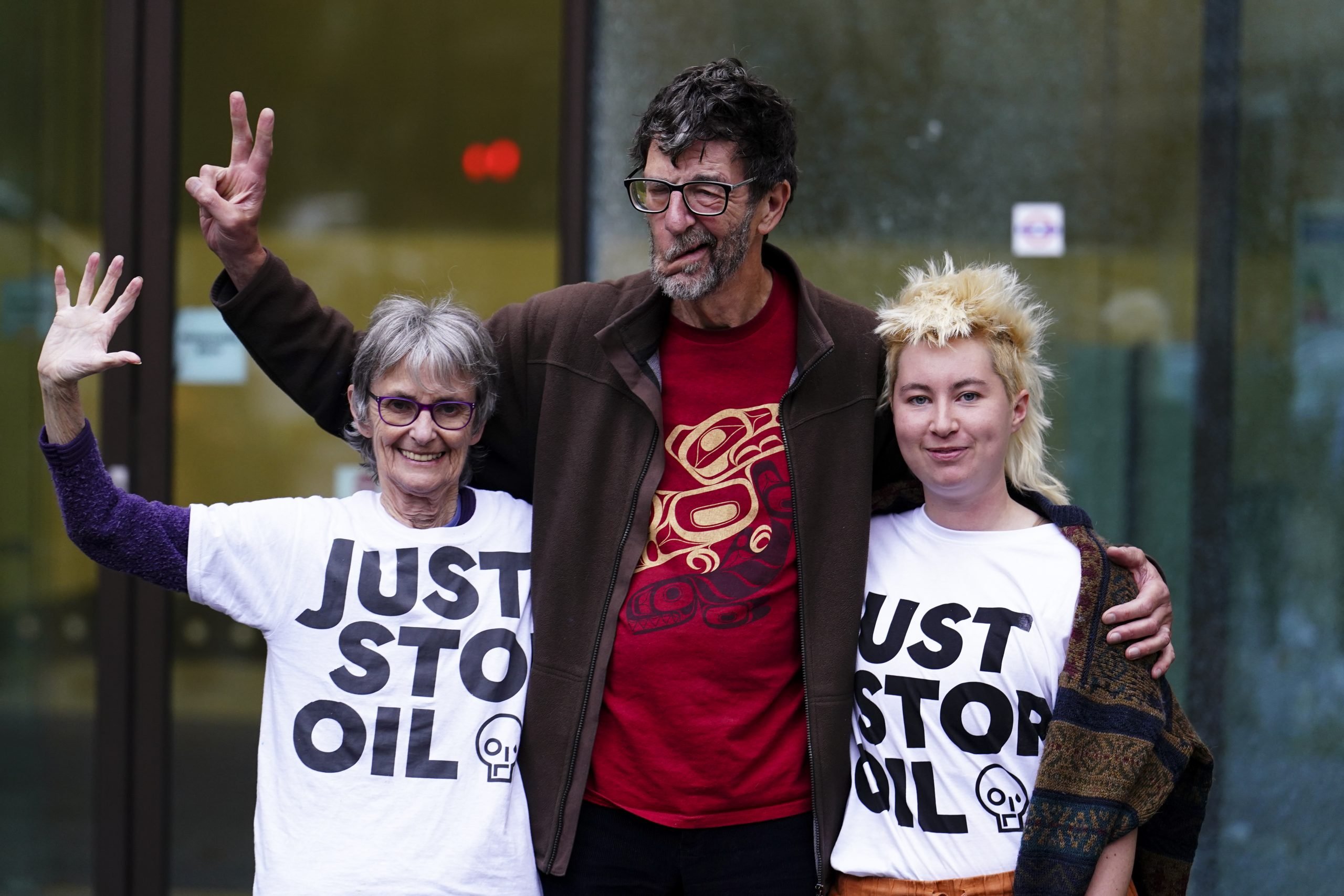 Three activists pose together, two wearing "Just Stop Oil" shirts, smiling and gesturing with peace signs in a public setting.