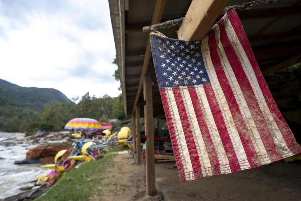 A dirt-stained American flag hangs from a wooden structure near colorful paddle boats by a river, with mountains in the background.