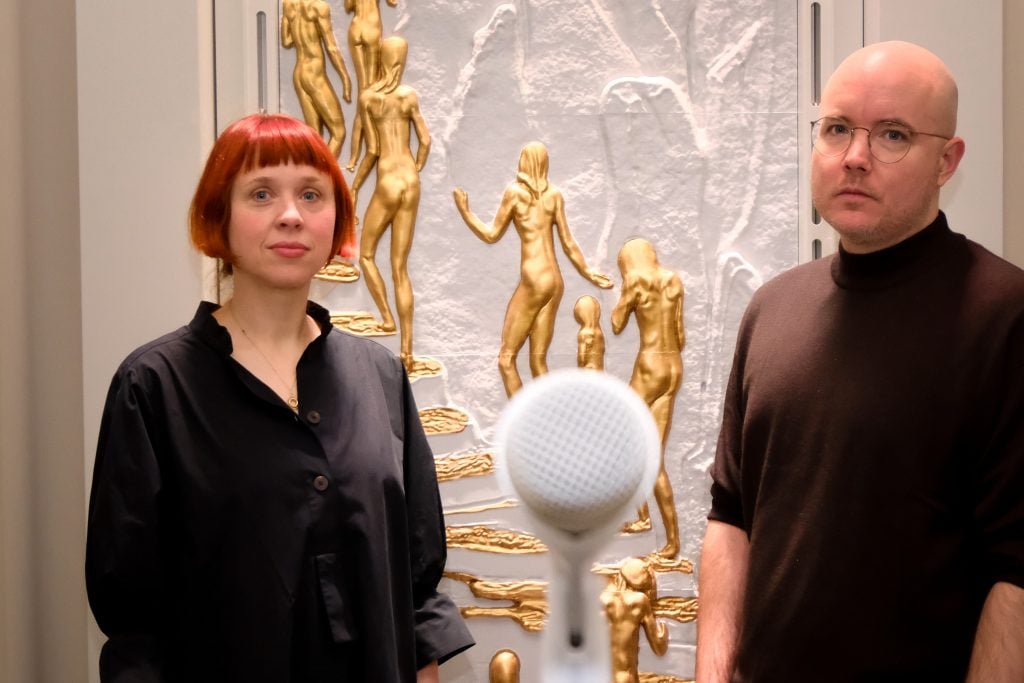 a man and a woman stand in front of an ornate sculptural artwork adorned with golden figures. there is a microphone between them in the foreground.