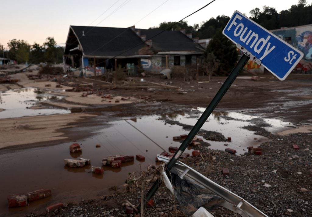 A tilted street sign reading "Foundy St" stands amidst muddy floodwaters, debris, and damaged buildings in a post-weather disasterscene.