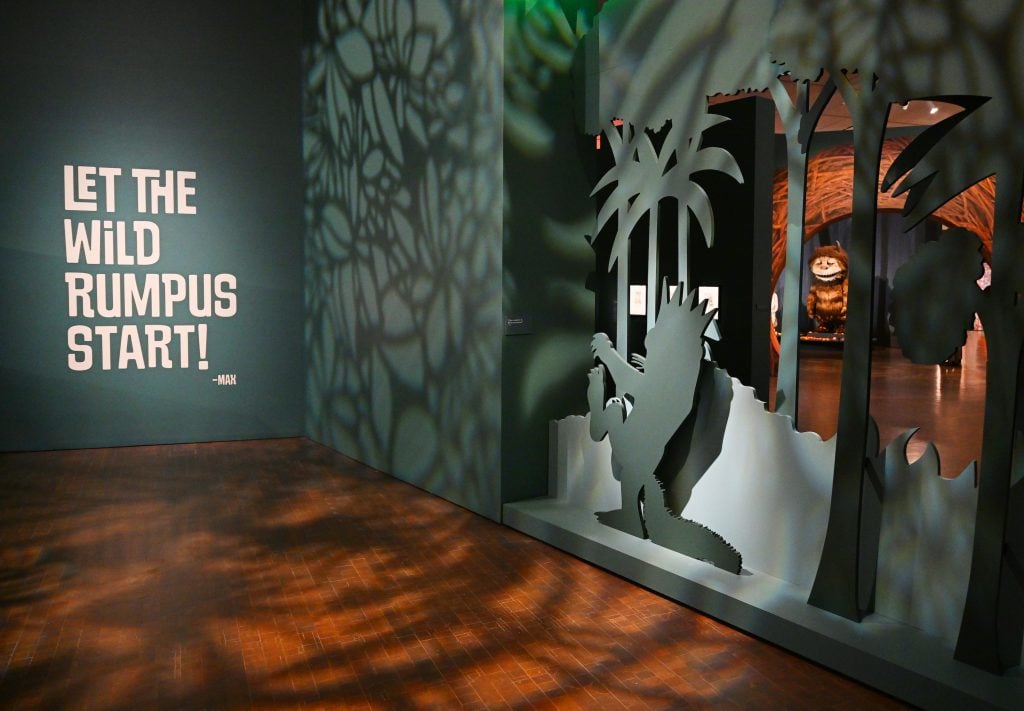 An exhibition display reads "LET THE WILD RUMPUS START!" alongside a shadowy cutout of a forest scene and a creature from Where the Wild Things Are in the background.