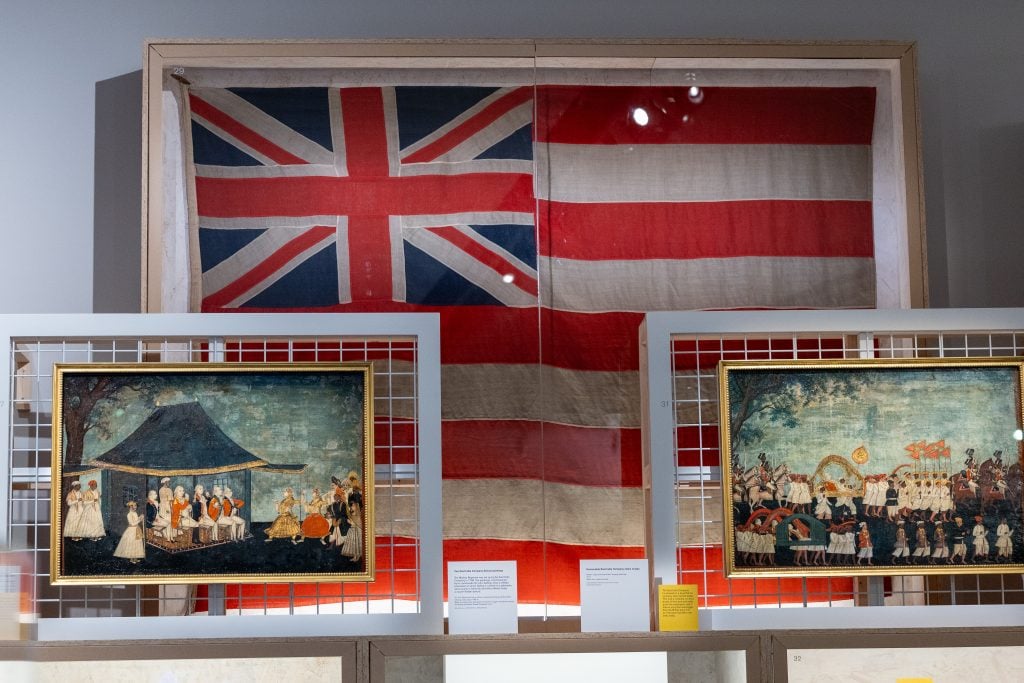 A historical display featuring a large Union Jack and East India Company flag, with framed paintings of British colonial scenes beneath.