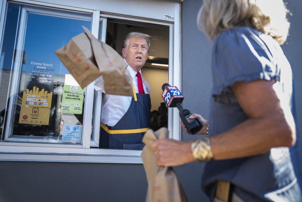 a white man holds brown paper bags outside of a drive-thru window