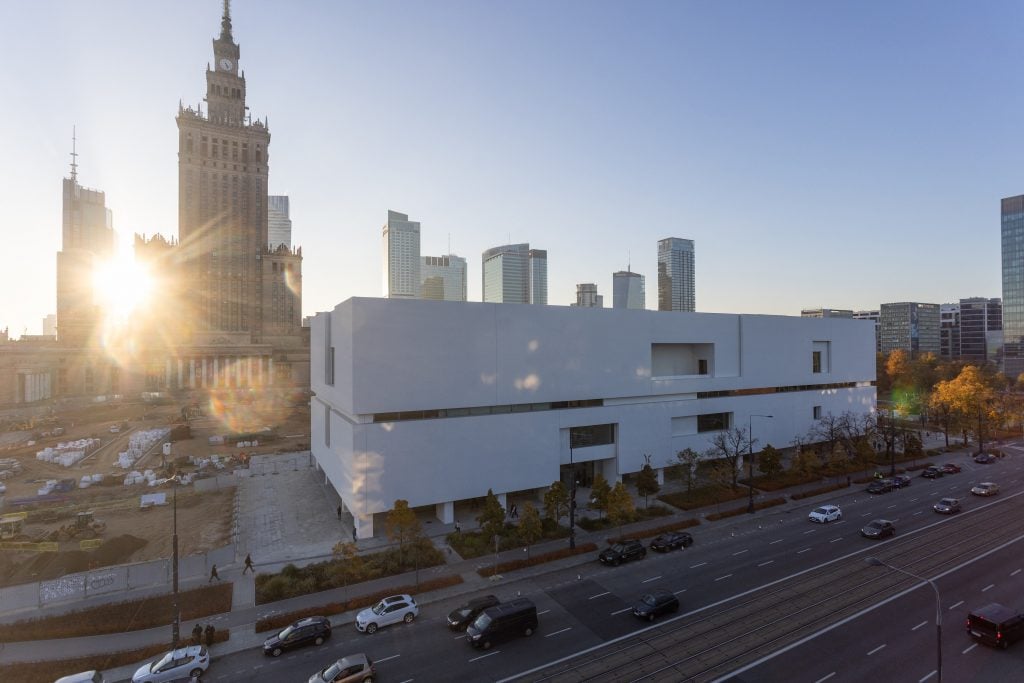 A white modern building in Warsaw with surrounding skyscrapers and the Palace of Culture and Science at sunrise in the background.