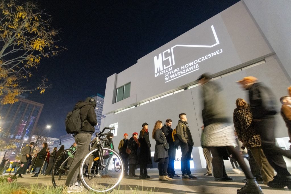 People stand in line outside the Museum of Modern Art in Warsaw at night, with a bicycle in the foreground.