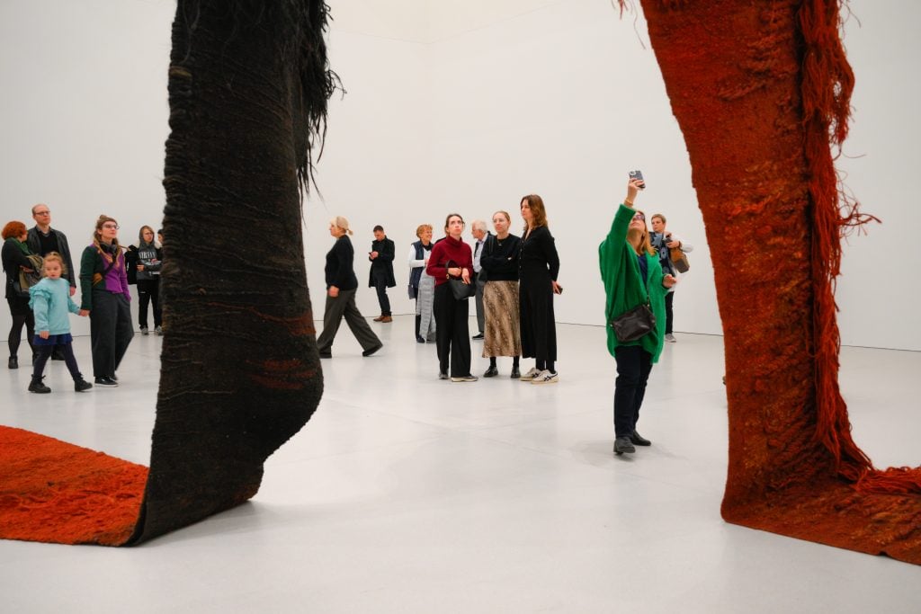 in a large white museum interior a small crowd of human figures are dwarfed by and look up at huge monumental hanging red fibrous textile in a deep red shade