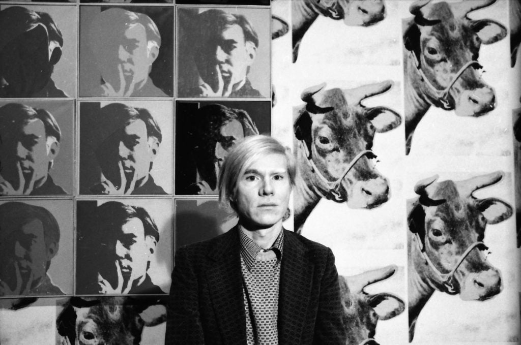 a black and white photo of artist andy warhol standing infront of his screen prints depicting cows