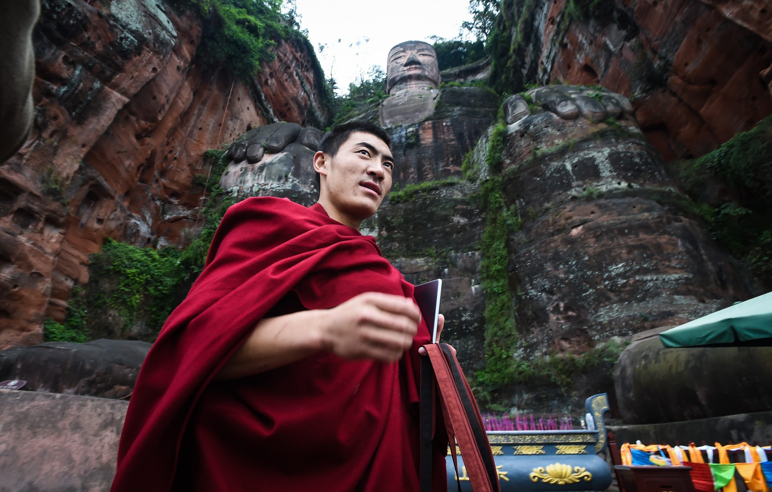 a monk wrapped in a red toga stands beneath the Leshan Buddha statue.