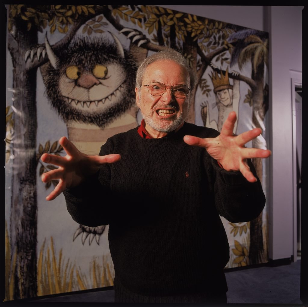 An elderly white man makes a growling face in front of life sized illustrations