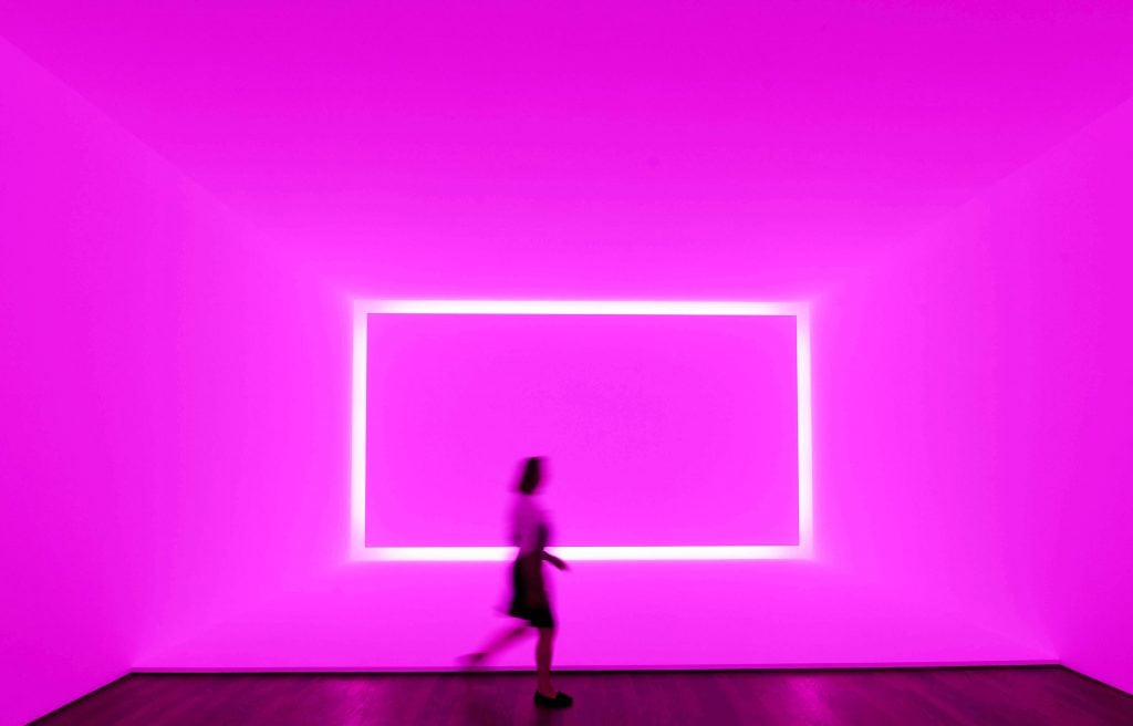 A person walks past a brightly lit, neon pink geometric installation with a glowing rectangular shape on the wall, creating a futuristic, immersive atmosphere