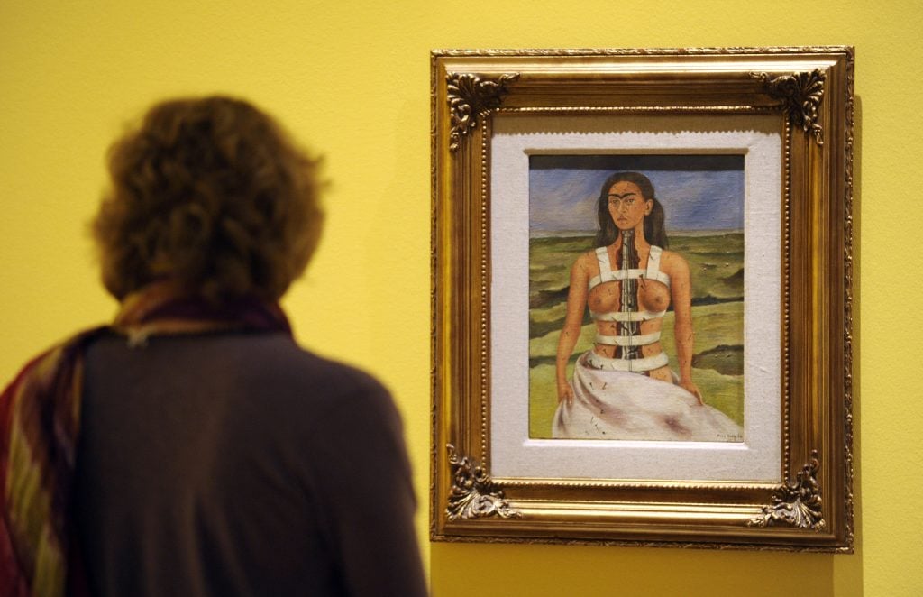 the back of a woman who is looking at a gold framed painting hung on a yellow wall. The painting is of a woman shown from the hips up who is wearing a metal brace and her torso appears ripped in two revealing a fractured Ionic column where her spine would be. She look sat us with a sad but stoic expression, tears are on her cheeks, there is a landscape behind