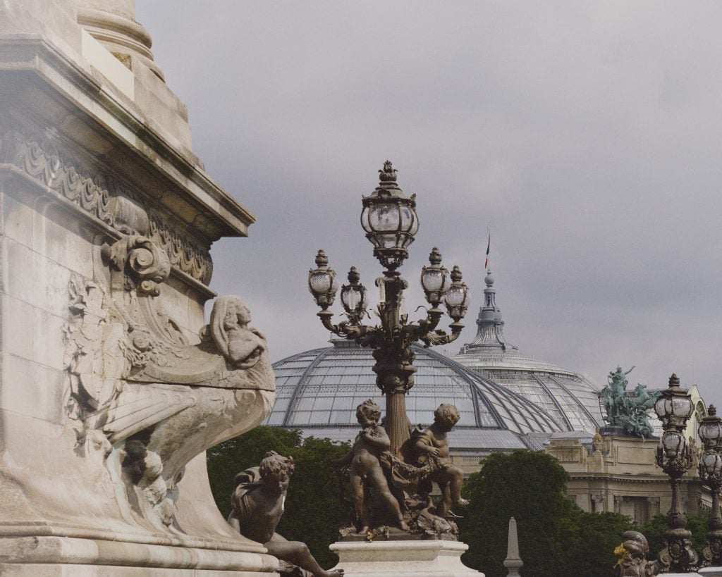 Decorative sculptures on bridge, with glass-domed building and French flag in background.