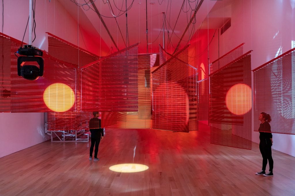 Two people dressed in black outfits standing under an installation of blinds lit in red lights in a gallery space