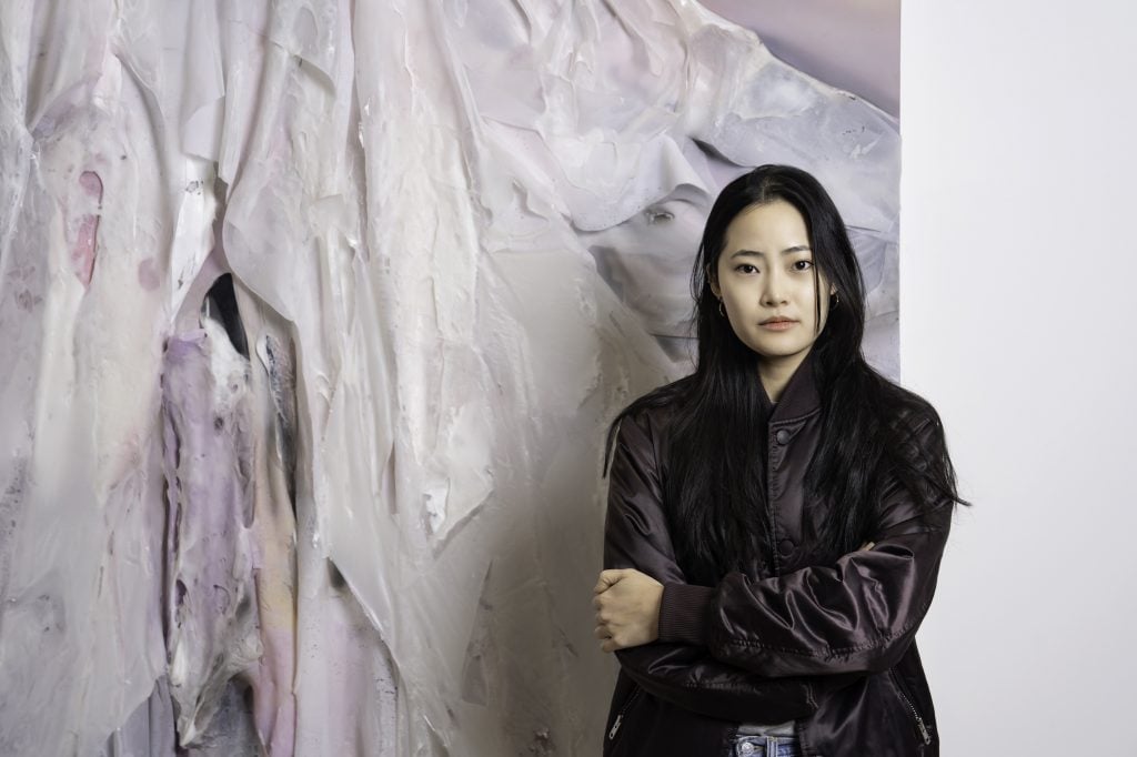 a young asian woman with black hair in a black leather jacket in front of an artwork with draping colors in hues of pink