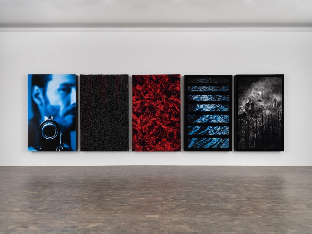 A series of five large panels displayed in a gallery setting. The first panel shows a close-up of Keanu Reeves, pointing a gun at the viewer, rendered in a blue-toned style. The second and third panels are abstract, with the middle one in vibrant red tones. The fourth panel features a series of blue lines resembling water, and the fifth panel shows a dark, atmospheric landscape with barbed wire in the foreground.