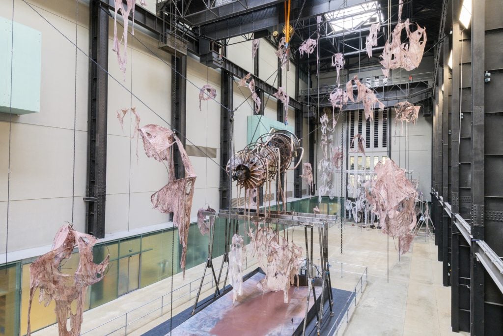 a very large hangar-like interior space filled by a strange installation that centers on a suspended metal turbine that drips danky pinky brown water down into a pool underneath, there are tufts of ragged fabric hanging all around