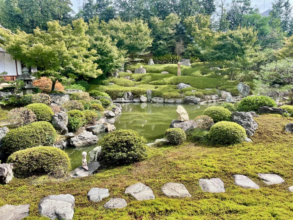 A Japanese garden full of green leaves and bushes, with sculptures installed around the pond in the middle. 