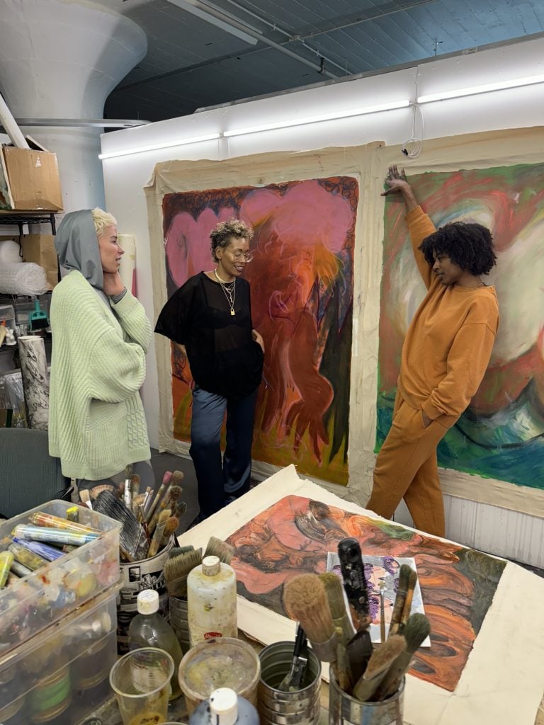 a group of three young women talk in an artist studio. paintings line the walls and brushes and paints are visible.