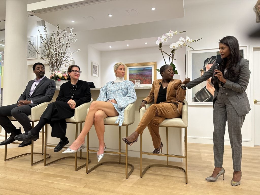 a group of four people, a man in a suit, a woman with glasses, a younger woman in pale blue, and another woman in a tan pansuit sit next to each other on a panel on raised chairs in a gallery space, a moderator addresses them